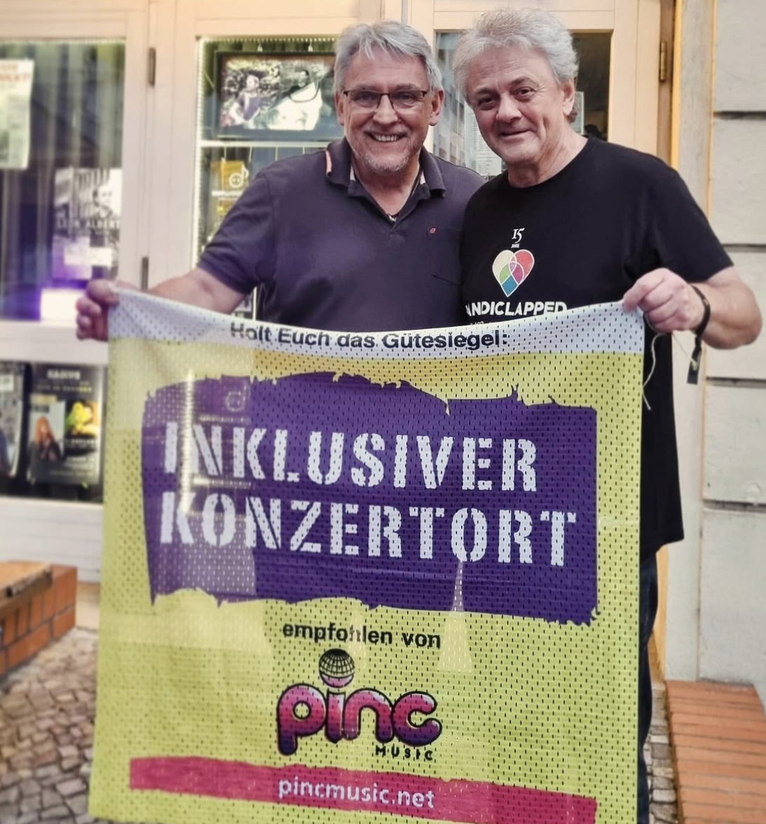 Bildbeschreibung: Buntfoto von zwei Männern die vor dem Eingang einer Kleinkunstbühne stehen, an dem einige Plakate hängen. Sie halten ein großes Stoffbanner hoch auf dem steht "holt Euch das Gütesiegel: Inklusiver Konzertort empfohlen von Pinc Music. pincmusic.net"