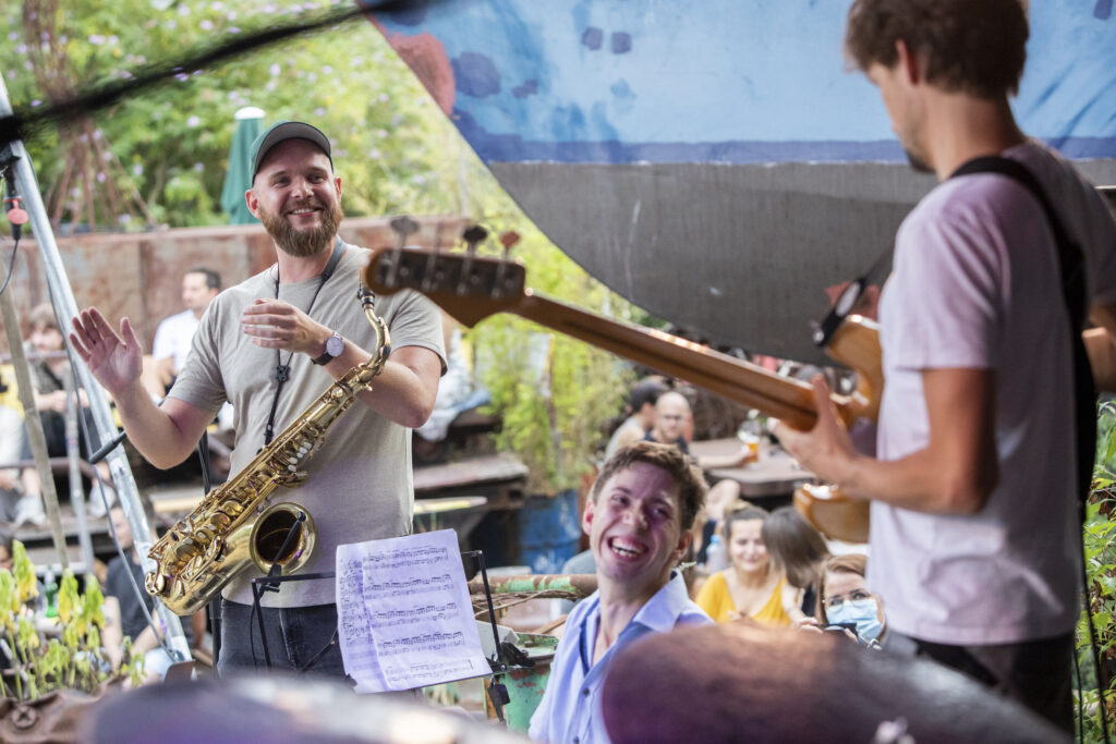 Buntfoto: Mehrere Musiker spielen gemeinsam draußen. Ein Mann mit Saxofon lächelt und gibt ein Zeichen mit der Hand. Ein anderer Musiker spielt Gitarre. Die Stimmung ist offen und fröhlich. Im Hintergrund sitzen Menschen und hören zu. Das Bild zeigt gemeinsames Musizieren und Austausch.