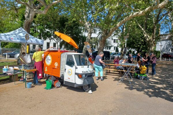 Ein Platz im Freien. Blauer Himmel ist zwischen den dicht belaubten Bäumen zu sehen. Unter Bäumen steht ein kleines dreirädriges Automobil in den Farben weiß und orange. Auf der Motorhaube steht die Aufschrift "Café-Treff". An der Seitenwand des Wagens leuchtet ein rundes Logo auf weißem Grund und gelb umrahmt: "Mein Lieblingsplatz". Eine Person mit gelbem T-Shirt und weinroter Schürze über einer blauen Jeans steht daneben. Auf dem Platz sind mehrere Personen, darunter eine im Rollstuhl, mehrere Festzeltgarnituren, ein orangefarbener Sonnenschirm und ein Zelt mit der Aufschrift "der Paritätische" zu sehen.