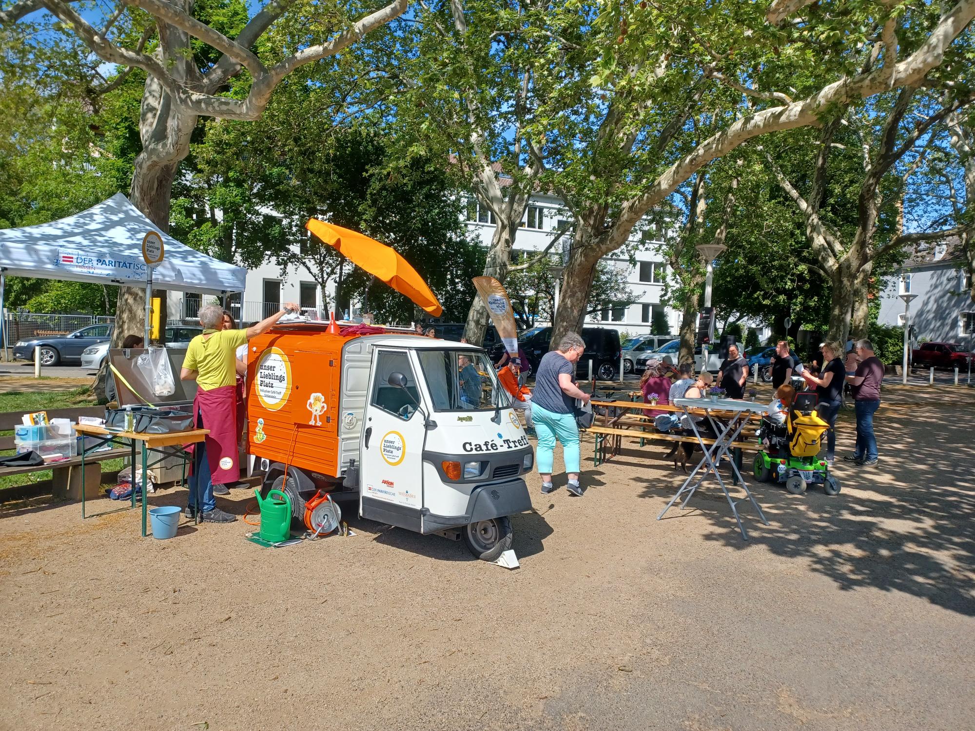Ein Platz im Freien. Blauer Himmel ist zwischen den dicht belaubten Bäumen zu sehen. Unter Bäumen steht ein kleines dreirädriges Automobil in den Farben weiß und orange. Auf der Motorhaube steht die Aufschrift "Café-Treff". An der Seitenwand des Wagens leuchtet ein rundes Logo auf weißem Grund und gelb umrahmt: "Mein Lieblingsplatz". Eine Person mit gelbem T-Shirt und weinroter Schürze über einer blauen Jeans steht daneben. Auf dem Platz sind mehrere Personen, darunter eine im Rollstuhl, mehrere Festzeltgarnituren, ein orangefarbener Sonnenschirm und ein Zelt mit der Aufschrift "der Paritätische" zu sehen.