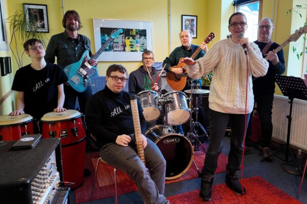Buntfoto von einer sechsköpfigen Band. Sie stehen in einem gelben Proberaum: Vorne rechts singt eine Frau mit Mikrofon, hinter ihr spielen zwei Männer Gitarre und Bass. In der Mitte sitzt ein junger Mann am Schlagzeug, davor ein weiterer mit einem Percussion-Instrument. Links stehen zwei Personen an Congas und E-Gitarre.