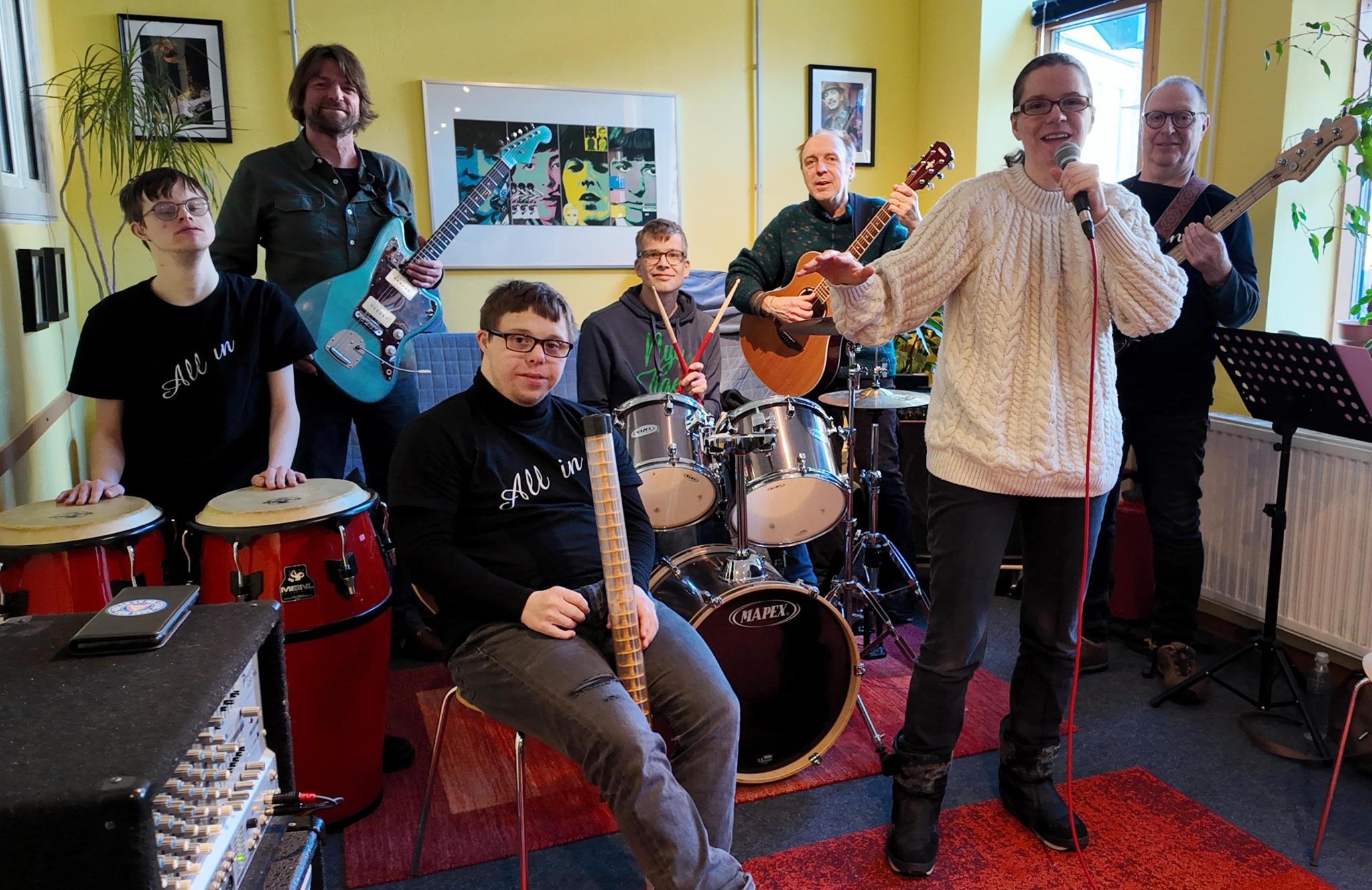 Buntfoto von einer sechsköpfigen Band. Sie stehen in einem gelben Proberaum: Vorne rechts singt eine Frau mit Mikrofon, hinter ihr spielen zwei Männer Gitarre und Bass. In der Mitte sitzt ein junger Mann am Schlagzeug, davor ein weiterer mit einem Percussion-Instrument. Links stehen zwei Personen an Congas und E-Gitarre.