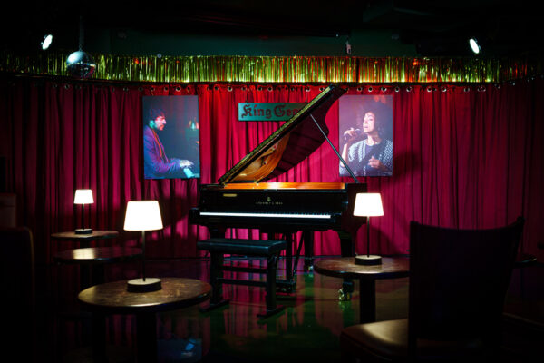 Buntfoto: Leere Bühne in einem kleinen Jazzclub: Ein schwarzer Flügel steht im Mittelpunkt vor roten Vorhängen, umgeben von kleinen runden Tischen mit gedämpften Tischlampen. An der Rückwand hängen Porträts von Musikern, die stimmungsvolle Beleuchtung schafft eine intime Clubatmosphäre.