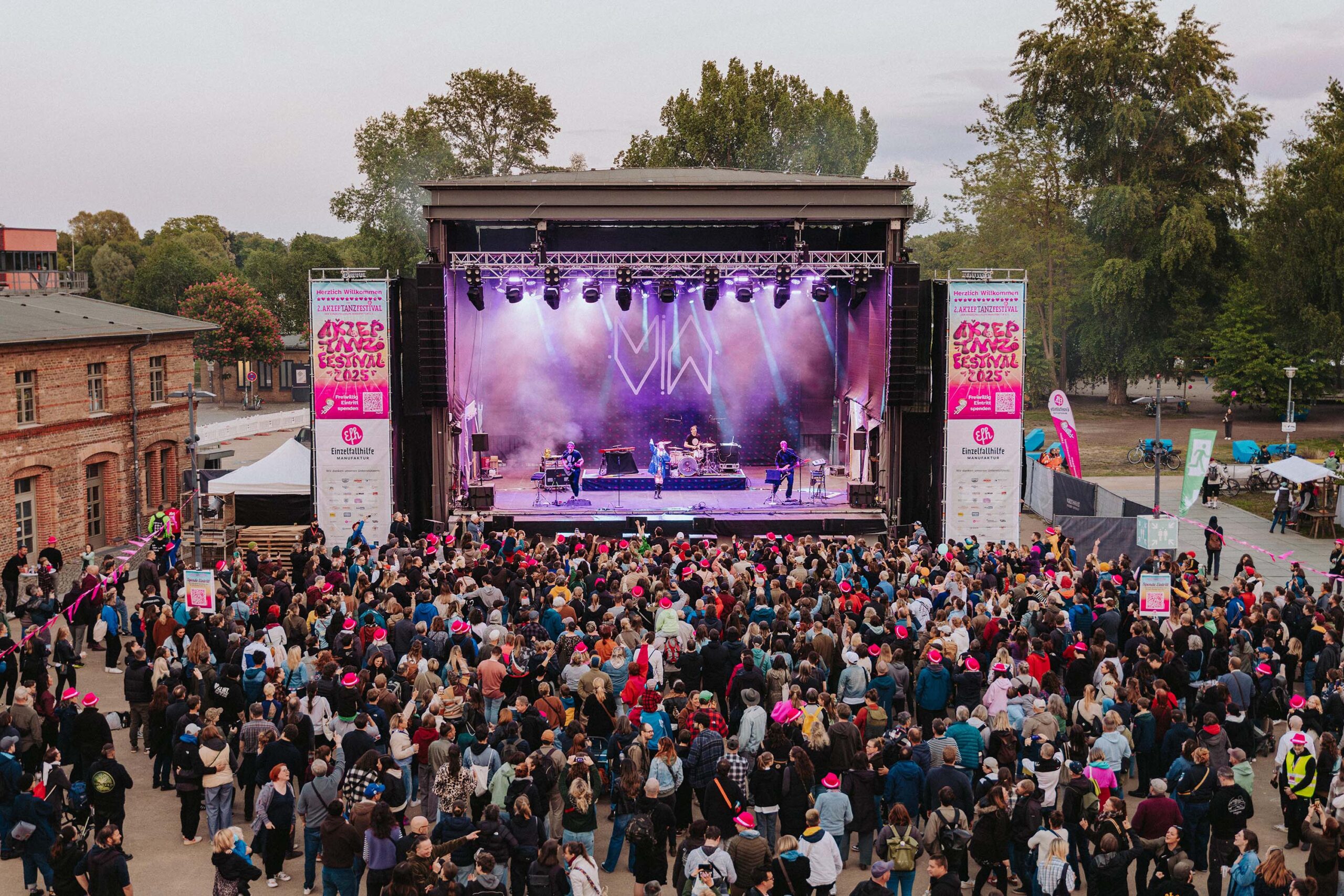 Buntfoto einer großen lila beleuchteten Bühne auf der eine Band auftritt davor ein großer Platz mit viel Publikum - hunderten von Menschen. IM Hintergrund hinter der Bühne sind Bäume.