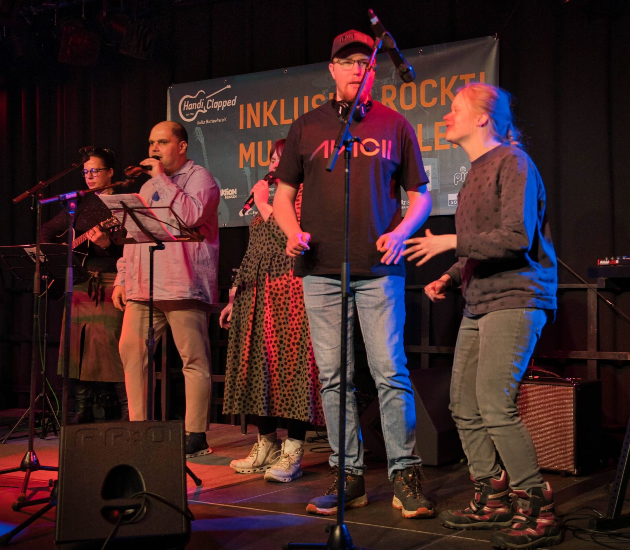 Buntfoto von zwei Sängerinen und zwei Sängern. Zwei singen in eigene Handmirkos, zwei zusammen in ein Standmikro. Im Hintergrund eine Bassistin.