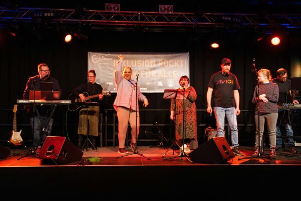 Buntfoto von sieben Musikern und Musikerinnen auf einer Bühne. Links und rechts jeweils ein Mann an einem Keyboard, dazwischen in der Mitte vier singende und eine Bassistin.