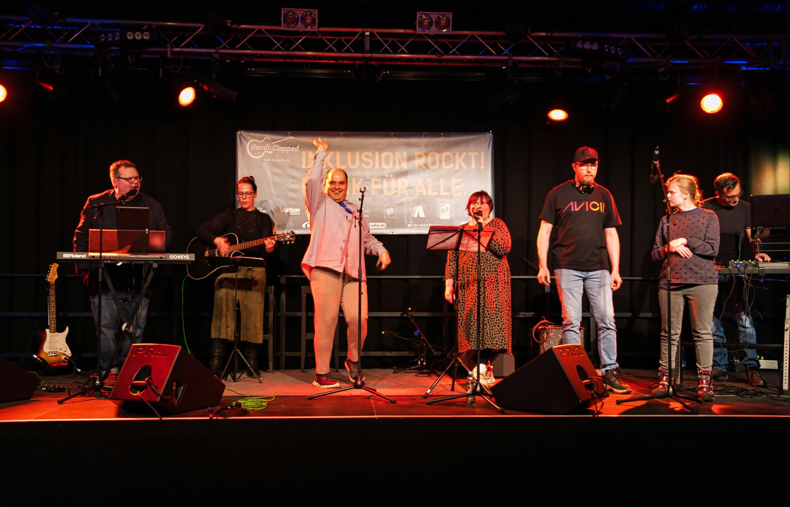 Buntfoto von sieben Musikern und Musikerinnen auf einer Bühne. Links und rechts jeweils ein Mann an einem Keyboard, dazwischen in der Mitte vier singende und eine Bassistin.
