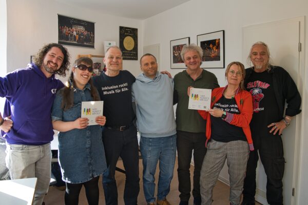 buntes Gruppenfoto von 2 Frauen und fünf Männern, in einem Büro, die eng zusammen stehen, zwei "Hier klingt's mir gut" Plaketten hochhalten und in die Kamera lächeln