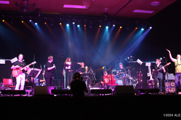 Konzertfotografie der POWERband auf einer Bühne bei einem Live-Auftritt. Das Bild zeigt eine vielköpfige Band während einer Performance in einer Veranstaltungshalle. Die Szene ist stimmungsvoll in violettes und blaues Bühnenlicht getaucht, wobei helle Scheinwerferstrahlen von oben auf die Musiker herabfallen.