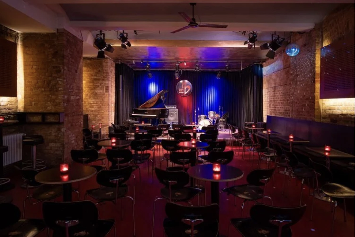 Buntfoto von einem Jazzclub im Keller, gediegene Atmosphäre mit ca 15 Tischen vor einer Bühne mit Flügel, Schlagzeug, Vorhang im Hintergrund und großem b-flat Logo davor hängend.