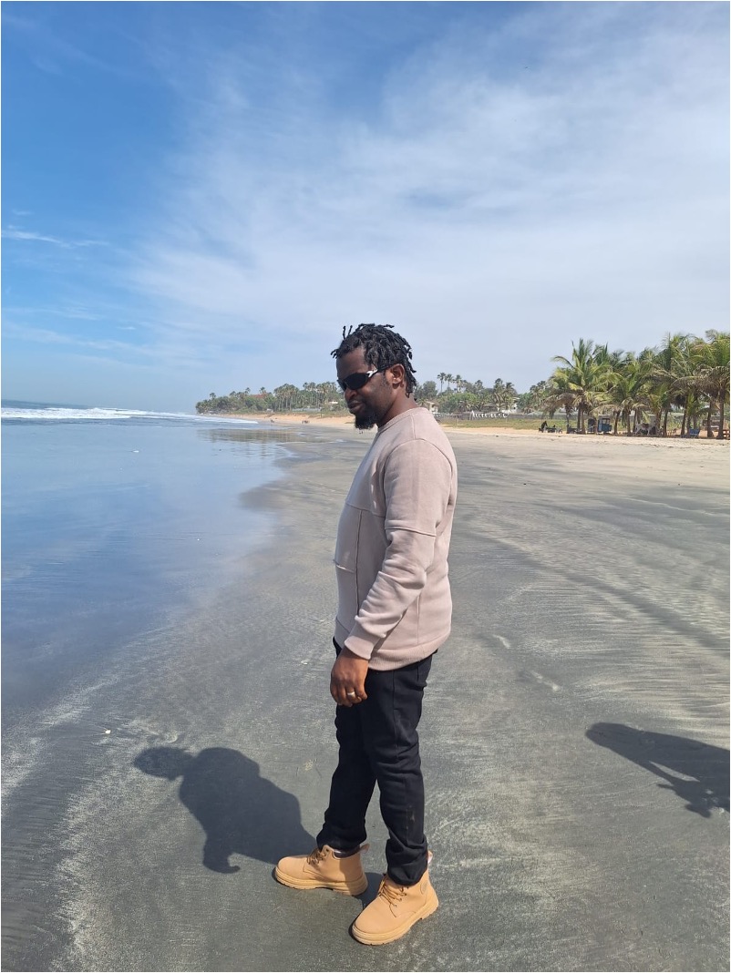 Ein Mann steht am Strand, links im bild das MEer, rechts der Strand, der Mann in der Mitte, er tr#gt einen beigen Pulli, eine dunkle lange Hose und helle Schuhe und eine Sonnenbrille.