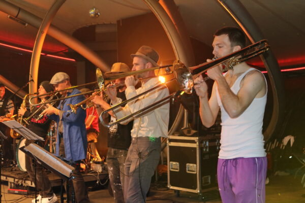 Buntfoto von sieben Mitgliedern einer Band, die Live auf der Bühne auftreten, zwei spielen Posaune, zwei Trompete und einer Saxophon. Sie sind von der Siete fotografiert. Fünf von ihnen tragen verschiedene Hüte und Kappen.