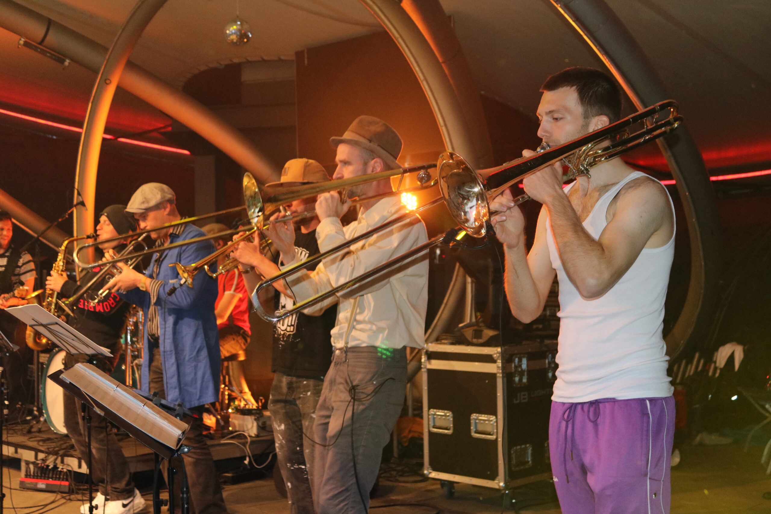 Buntfoto von sieben Mitgliedern einer Band, die Live auf der Bühne auftreten, zwei spielen Posaune, zwei Trompete und einer Saxophon. Sie sind von der Siete fotografiert. Fünf von ihnen tragen verschiedene Hüte und Kappen.