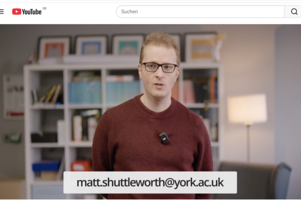 Bildschirmfoto von einem YouTubeVideo in dem ein junger mann im roten Pulli und mit Pulli in die Kamera psricht. Er staht vor einem modernen Bücherregal
