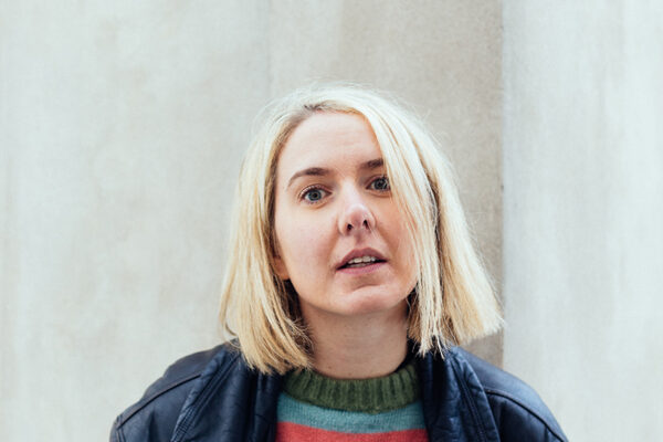 Buntfoto Portrait von Kopf und Oberkörper einer Frau im gestreiften Pulli und dunkler Jacke vor einer hellen Wand