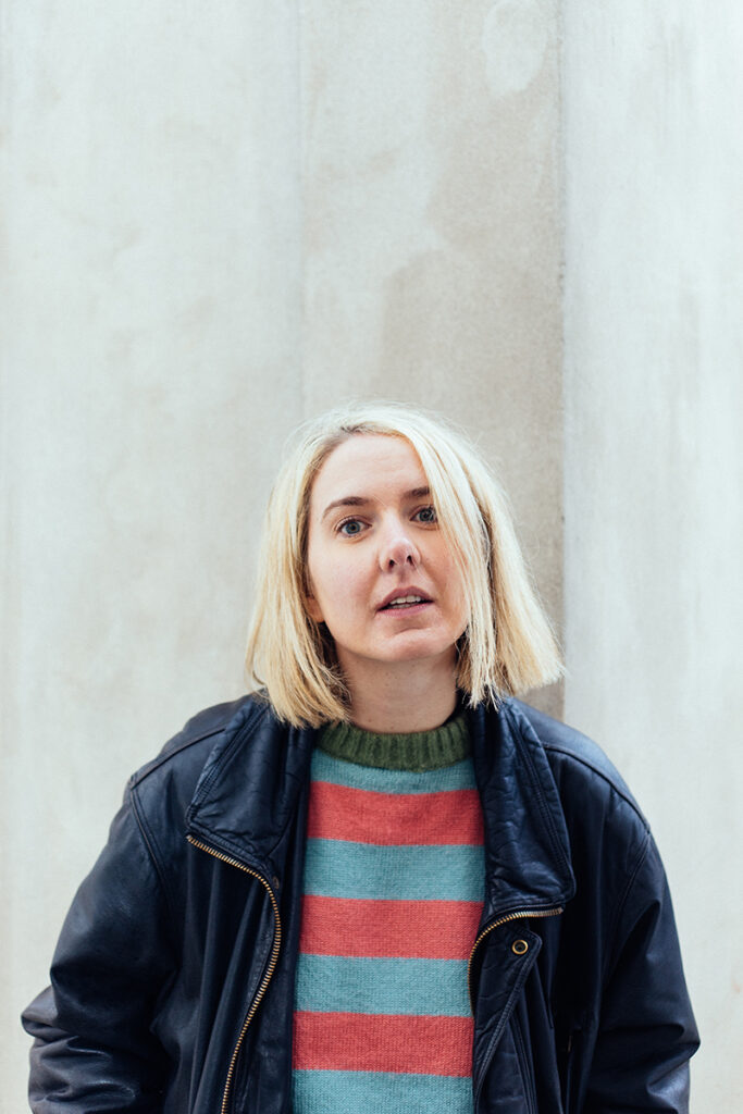 Buntfoto Portrait von Kopf und Oberkörper einer Frau im gestreiften Pulli und dunkler Jacke vor einer hellen Wand