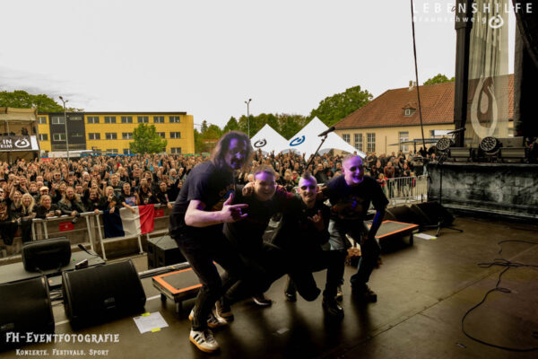 Buntfoto von einer Band aus vier Männern, die auf einer Bühne zum Gruppenfoto posiert. Im Hintergrund ein Marktplatz ranvoll gefüllt mit einer Menschenmenge. Oben rechts ist das Logo der Lebenshilfe Braunschweig über dem Foto, unten links "FH-Eventfotografie - Konzerte Festivals Sport"