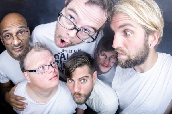 Gruppenfoto von fünf Männern von oben fotografiert, alle tragen weiße E-Shirts und schneiden Grimassen.