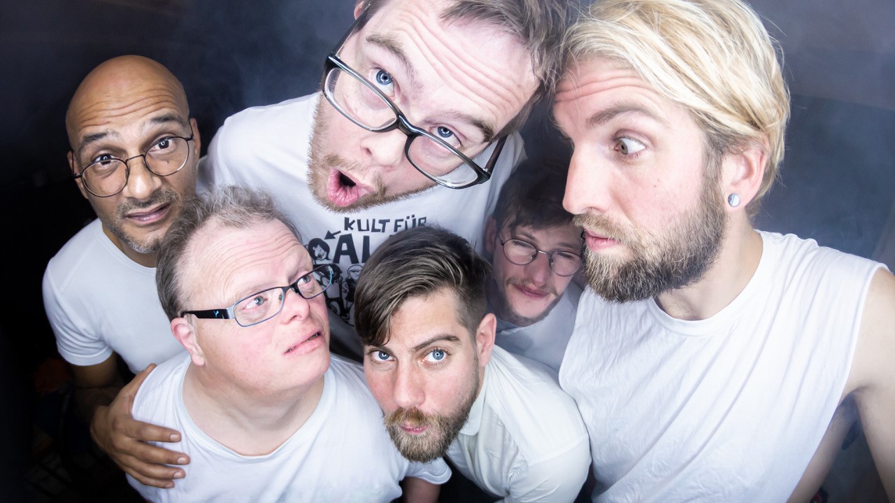 Gruppenfoto von fünf Männern von oben fotografiert, alle tragen weiße E-Shirts und schneiden Grimassen.