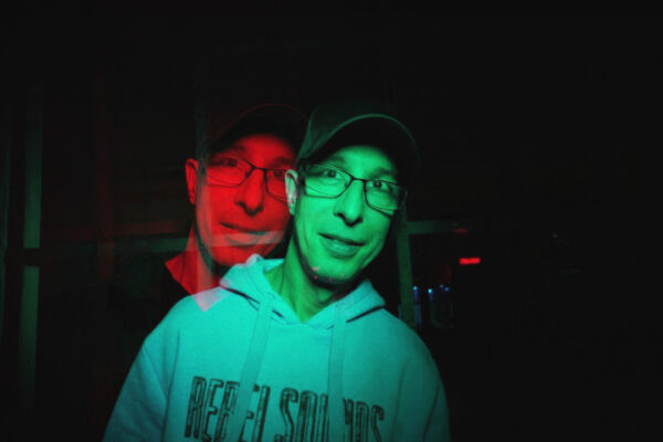Eine Person mit Brille und dunklem Basecap trägt einen hellen Kapuzenpullover und blickt in die Kamera. Durch einen Doppelbelichtungseffekt und rot-grün aufgeteilte Beleuchtung erscheint das Gesicht zweimal leicht versetzt. Der Hintergrund ist dunkel und nur schwach erkennbar.