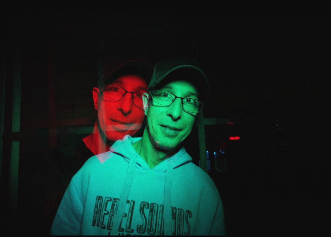 Eine Person mit Brille und dunklem Basecap trägt einen hellen Kapuzenpullover und blickt in die Kamera. Durch einen Doppelbelichtungseffekt und rot-grün aufgeteilte Beleuchtung erscheint das Gesicht zweimal leicht versetzt. Der Hintergrund ist dunkel und nur schwach erkennbar.
