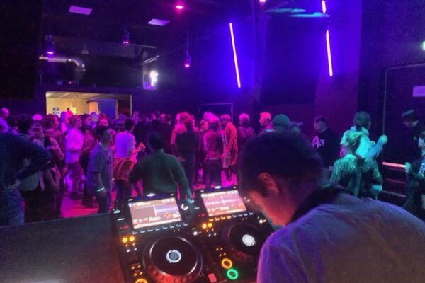 In einem lila-blau ausgeleuchteten Raum steht ein DJ am Pult. Im gefüllten Raum sind ca. 50 tanzende Menschen zu sehen.