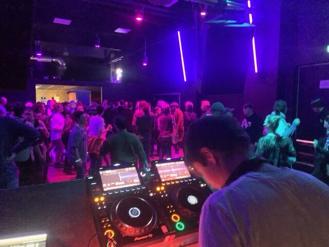 In einem lila-blau ausgeleuchteten Raum steht ein DJ am Pult. Im gefüllten Raum sind ca. 50 tanzende Menschen zu sehen.