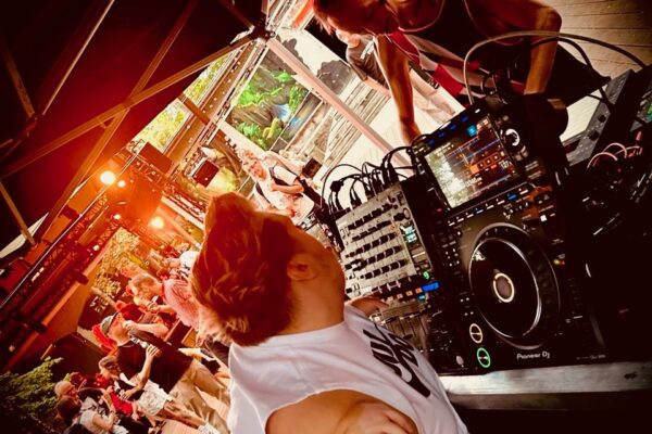 Zwei DJs bei einem Open-Air Event. zwischen ihnen stehen Mischpult und Player. Im Hintergrund tanzen Menschen. Es ist eine schräge Kameraperspektive gewählt.