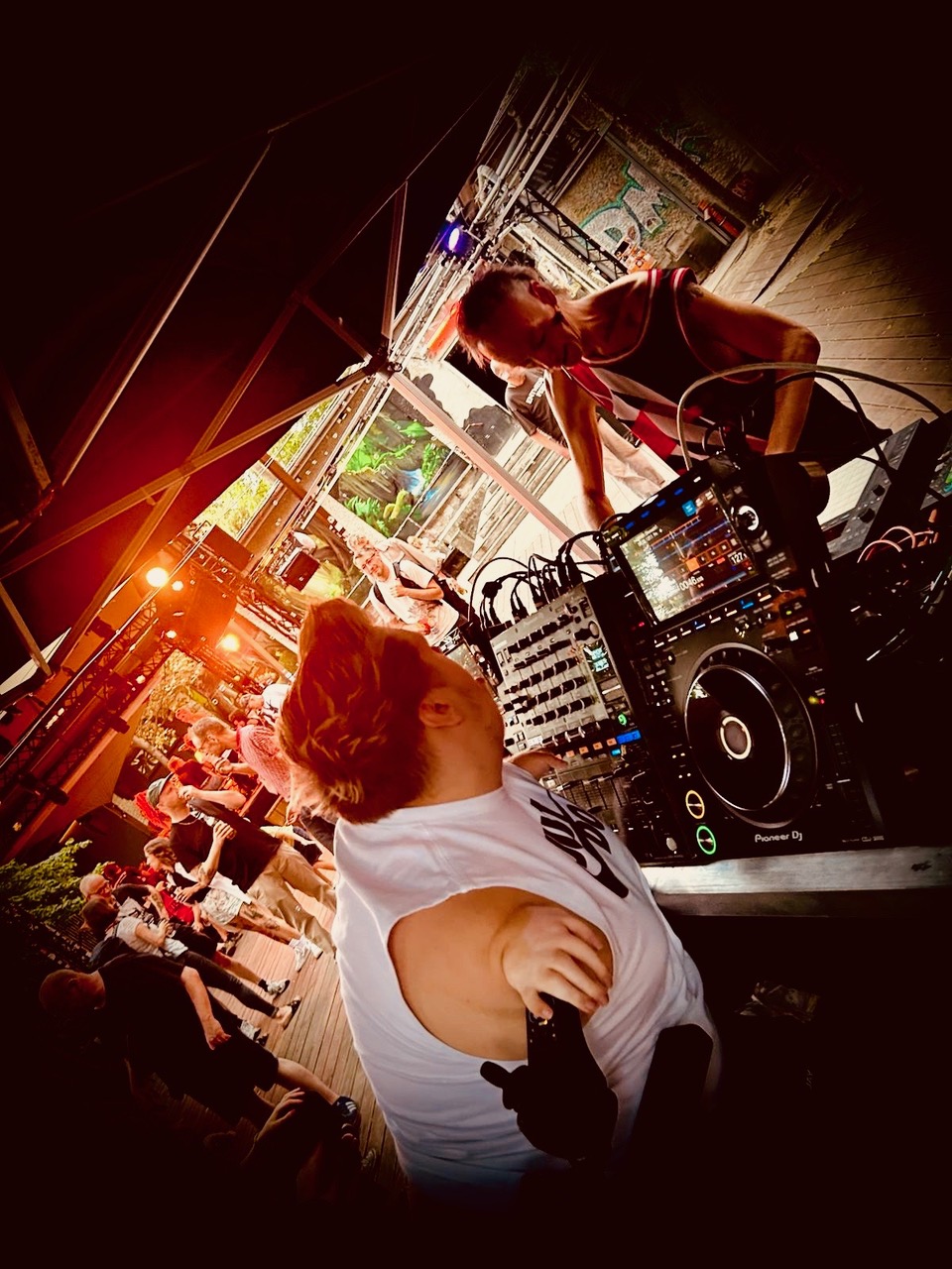Zwei DJs bei einem Open-Air Event. zwischen ihnen stehen Mischpult und Player. Im Hintergrund tanzen Menschen. Es ist eine schräge Kameraperspektive gewählt.