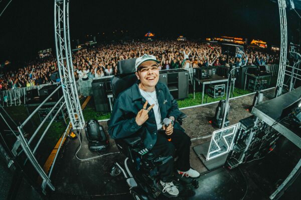 DJ LUCA VEDA sitzt lächelnd auf einer Festivalbühne im Rollstuhl, hält ein Mikrofon, formt mit der anderen Hand ein Peace-Zeichen und blickt von der Bühne auf das feiernde Publikum im Hintergrund.