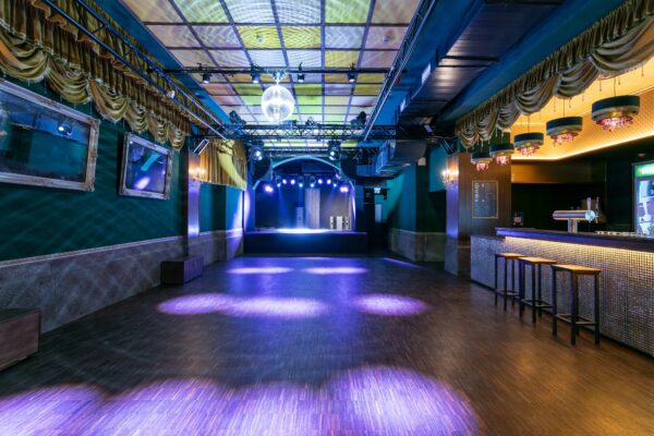Innenansicht eines leeren, stimmungsvollen Nachtclubs mit einer zentralen Tanzfläche und einer Bühne am Ende. Links sind gerahmte Spiegel an einer grünen Wand, rechts eine Bar mit Hockern und die Decke ist mit Lichteffekten, Vorhängen und einer Discokugel geschmückt.