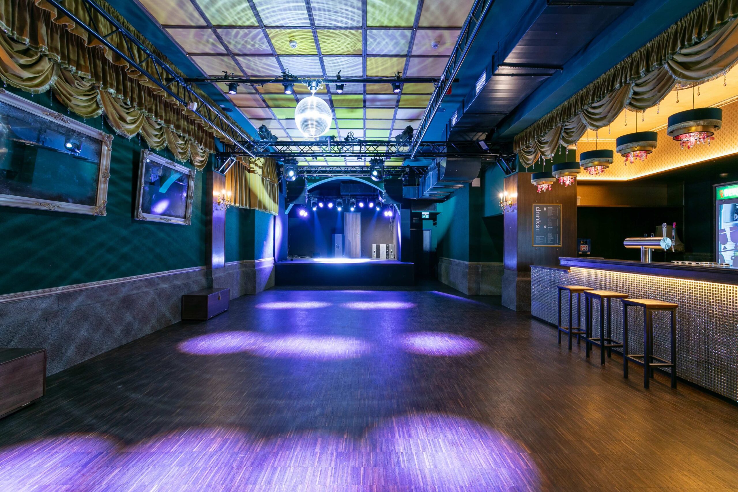 Innenansicht eines leeren, stimmungsvollen Nachtclubs mit einer zentralen Tanzfläche und einer Bühne am Ende. Links sind gerahmte Spiegel an einer grünen Wand, rechts eine Bar mit Hockern und die Decke ist mit Lichteffekten, Vorhängen und einer Discokugel geschmückt.