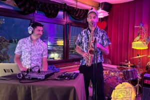 Ein Saxophonist und ein Mann neben ihm, der einen Kopfhörer  trägt und ein Mischpult bedient.  Alles live auf einer Studiobühne. Im hinbtergund ein großes Fenster mit einem Stadtpanorama.
