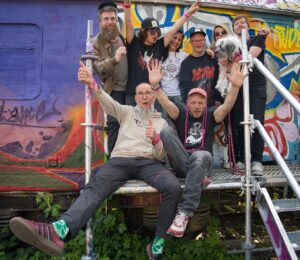 Gruppefoto von acht in die Kamera jubelnden menshen und einem Hund, vor einer Graffity Wand