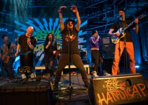 Zwei Musikerinnen die Singen und drei Musiker, zwei Sänger und ein Gitarrist live auf der Bühne. Dahinter buntes Bühnenbild