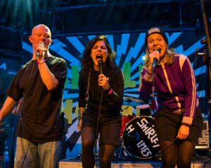 Ein Mann und zwei Frauen singen live auf der Bühne in drei Handmikrofone die sie halten. Dahinter buntes Bühnenbild