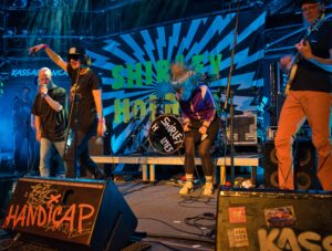 Drei Msuiker und eine Msuikerin live auf der Bühne. Im Vordergrund fette Lautsprecher. Auf einem Steht HandyCap. Dahinter buntes Bühnenbild
